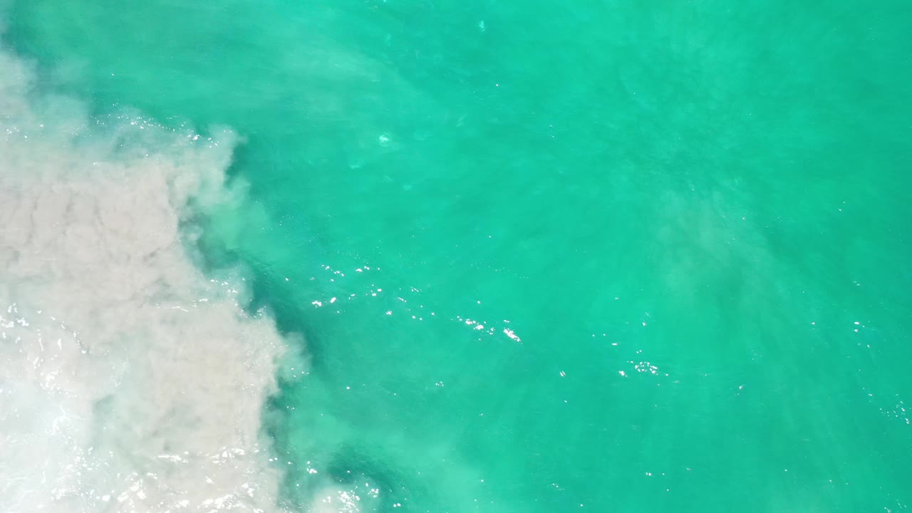 Overhead Los Cabos water frame with pale foam expanding from the lower left and small sunlight reflections across the blue-green surface, no visible people.