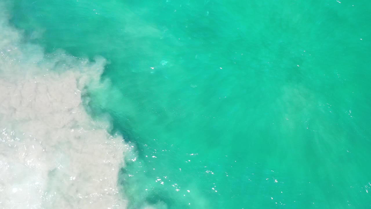 Aerial view over turquoise Los Cabos water, with a bright white surf mass on the left and open clear water across the right, no visible people.