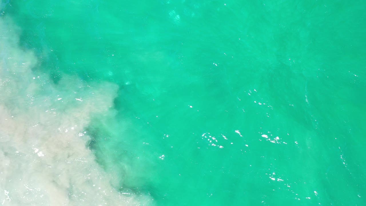 Overhead view in Los Cabos, Mexico, showing turquoise ocean water beside foamy pale coastal wash at the left edge, with no visible people.