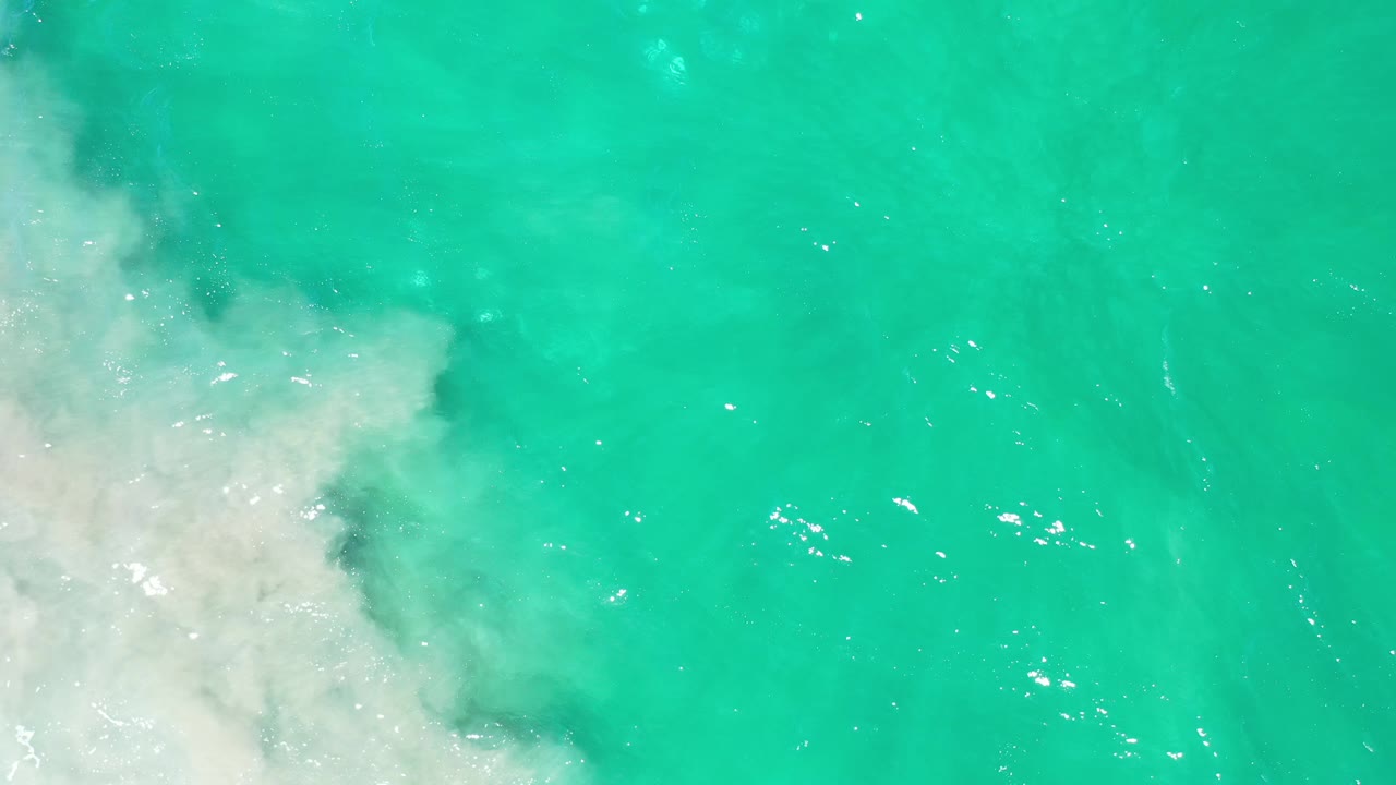 Overhead Los Cabos water view with a larger white foam patch pushing in from the lower left and open turquoise water across the right, with no visible people.