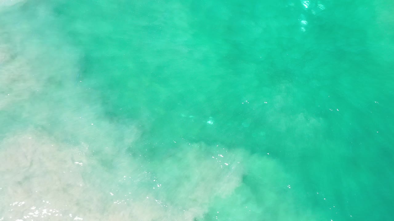 Overhead view of turquoise water washing across pale sand in Los Cabos, Mexico, with soft foam at the lower left and no visible people.