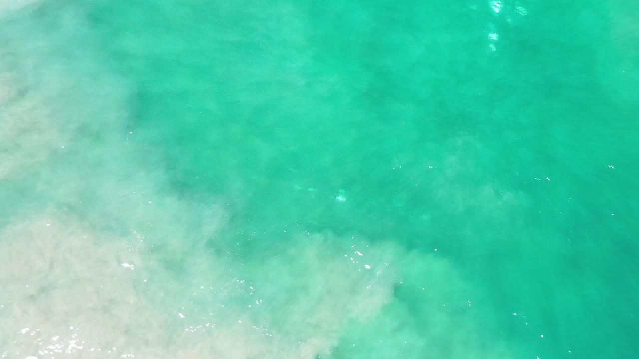 Aerial view of green turquoise ocean crossing sandy shallows in Los Cabos, Mexico, with a broad foam band near the bottom and no visible people.