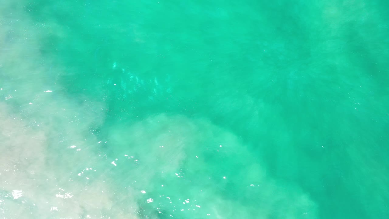 Top-down aerial frame of turquoise green water in Los Cabos, Mexico, with pale surf and sand-colored shallows near the lower left, no visible people.