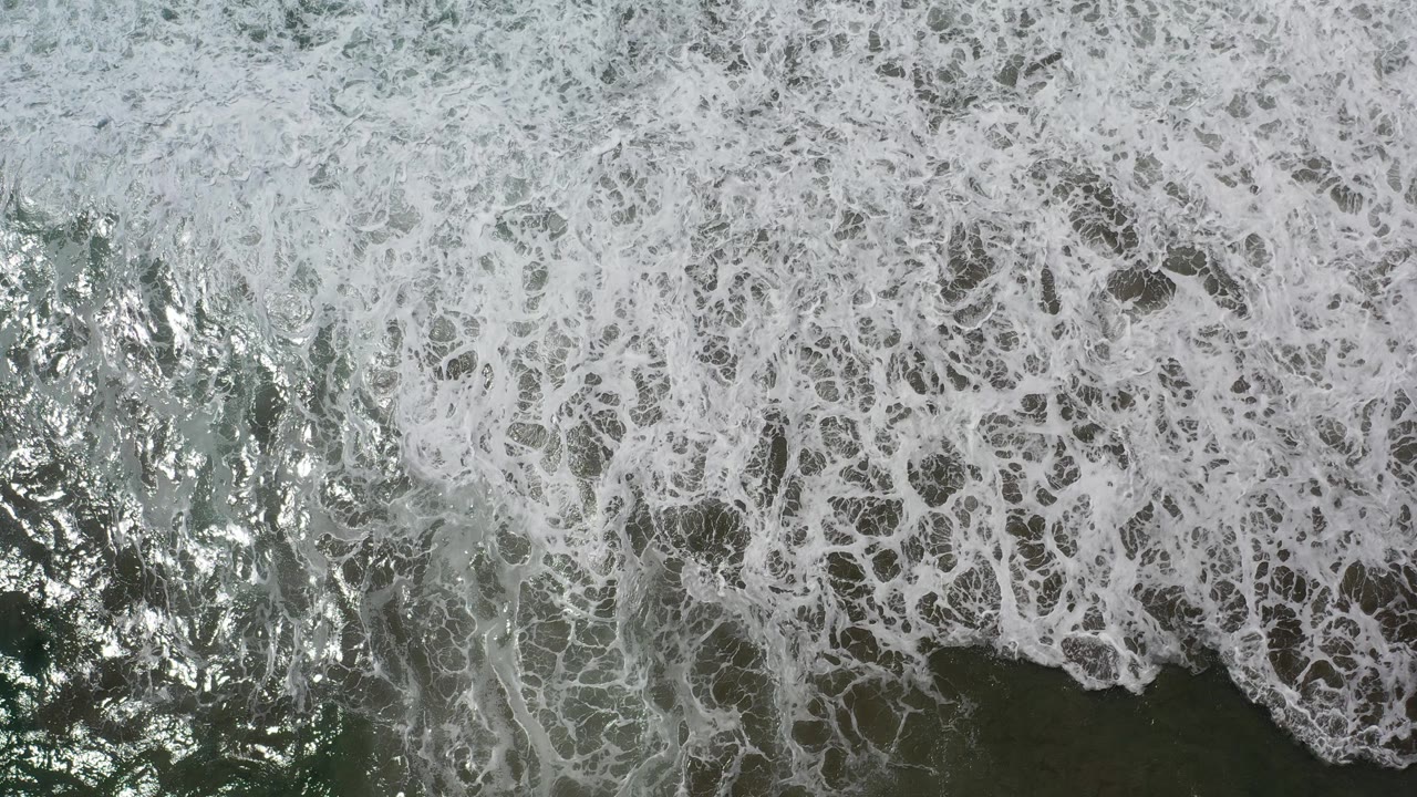 Overhead view of rough sea water in Los Cabos, Mexico, with dense white foam spreading across dark green-gray water and no visible people.