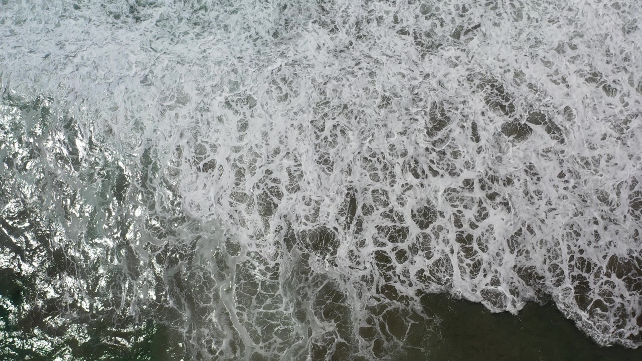 Overhead Los Cabos ocean frame with foamy white surf curling around a darker water patch near the lower edge and no visible people.