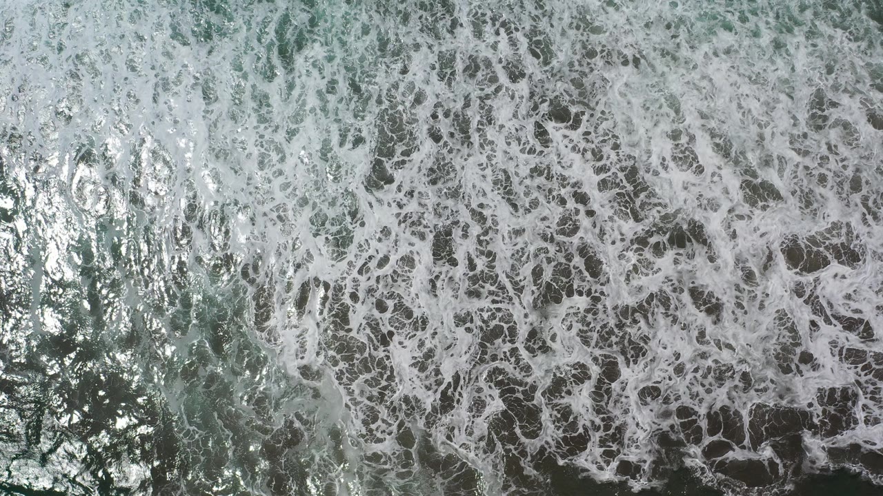 Overhead frame of white surf foam spreading across green ocean water in Los Cabos, Mexico, with bright reflections and no visible people.