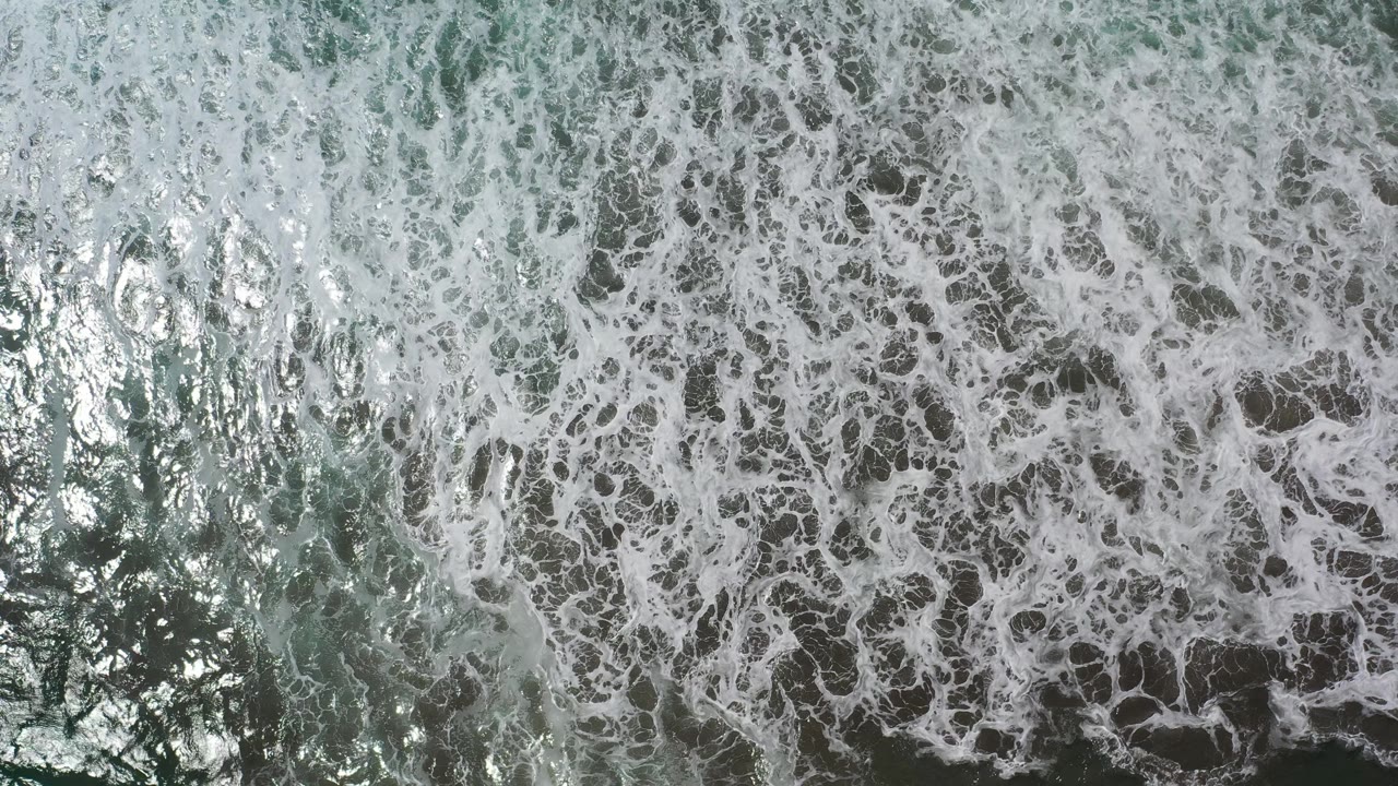 White foam patterns cover most of the Los Cabos ocean surface while green reflective water shows through near the lower left, with no visible people.