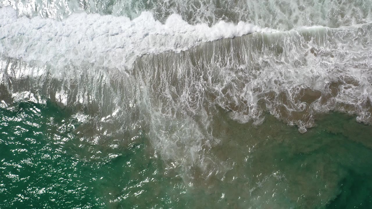 Overhead view of blue-green water in Los Cabos with a white surf band crossing the top edge, darker submerged shapes below, and no visible people.