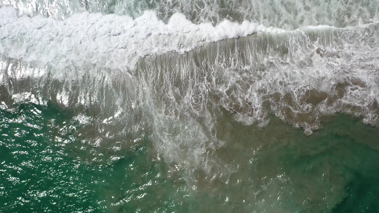 Aerial Los Cabos frame with white foam pushing farther down over translucent green water and mottled brown submerged areas, with no visible people.
