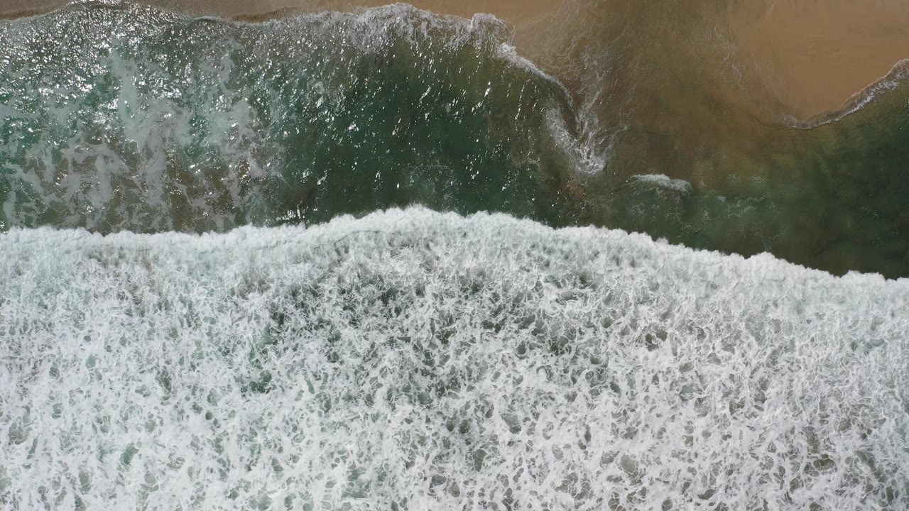 Overhead view of white surf breaking across green water and tan sand in Los Cabos, Mexico, with no visible people.