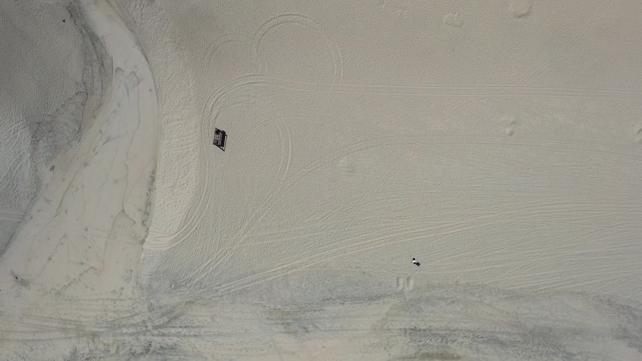 Straight-down aerial view of pale Los Cabos sand with curved tire tracks, a drainage edge at left, and a small visible person near the lower part of the frame.