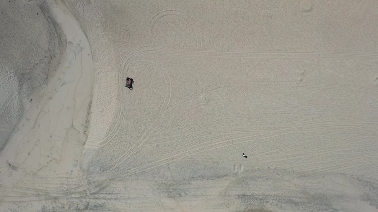 Aerial top-down frame over Los Cabos beach sand, showing sweeping tire tracks, a dark channel along the left side, and a tiny visible person or object on the sand.