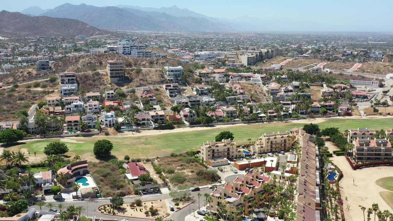 Aerial frame in Los Cabos showing homes and apartment blocks below dry hills, with a broad green strip crossing the foreground and distant mountains under bright sky.