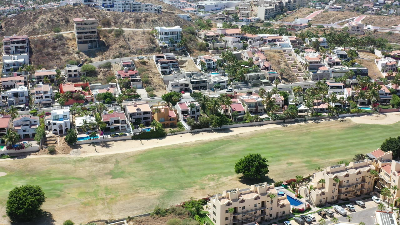 Los Cabos aerial view with the fairway occupying the lower frame, a row of villas and palms along a road, scattered hillside construction, and no visible people.