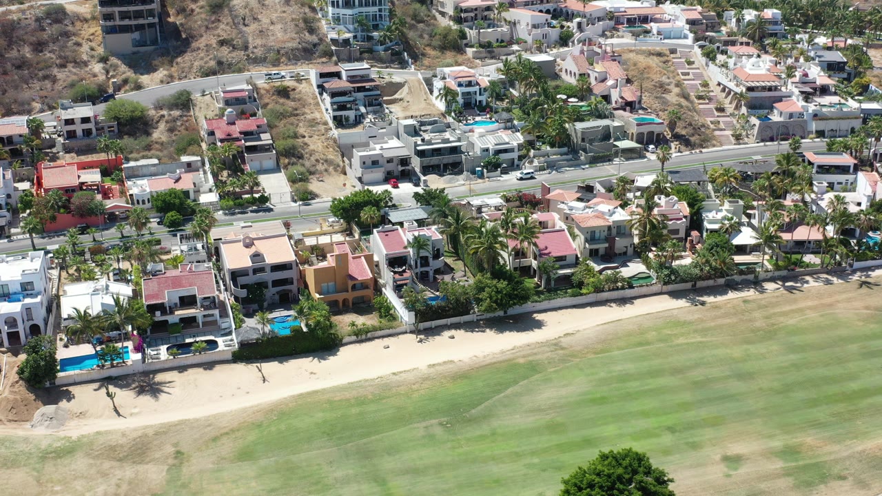 Aerial view of beachside homes, pools, road, and dry hillside.