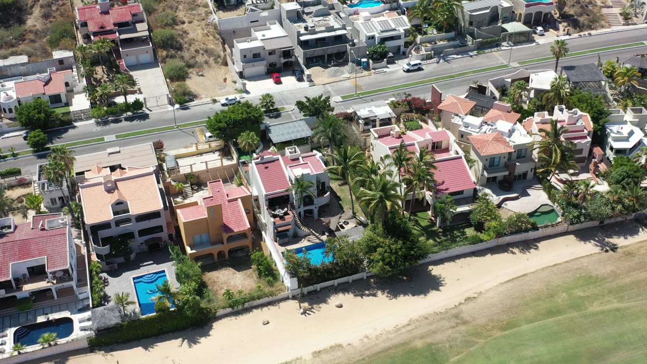 Aerial view of beachfront houses, pools, palms, and a road.