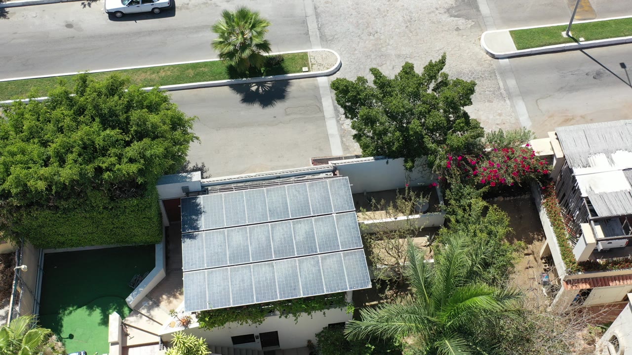 Early overhead frame in Los Cabos, Mexico, showing a quiet road above walled residential properties, a metal-roofed carport, palms, and no visible people.