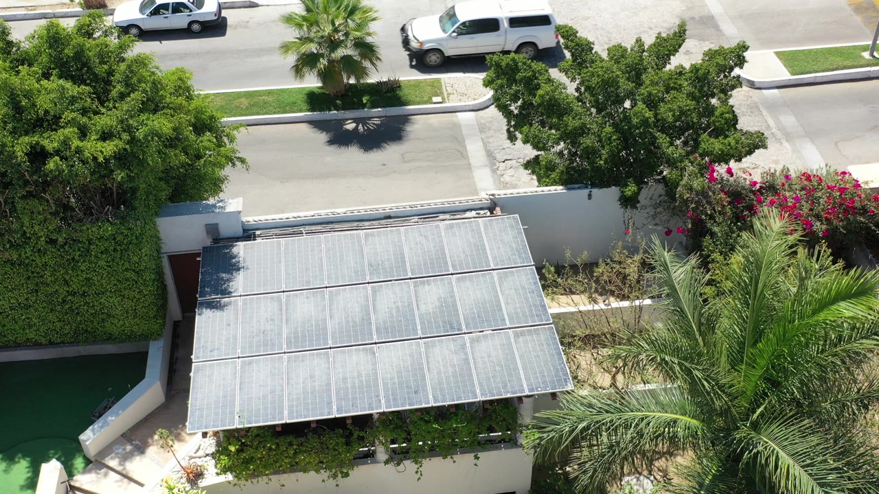 Opening aerial frame in Los Cabos, Mexico, showing a white car on the street above a walled home, with a metal roof, hedges, palm fronds, and no visible people.