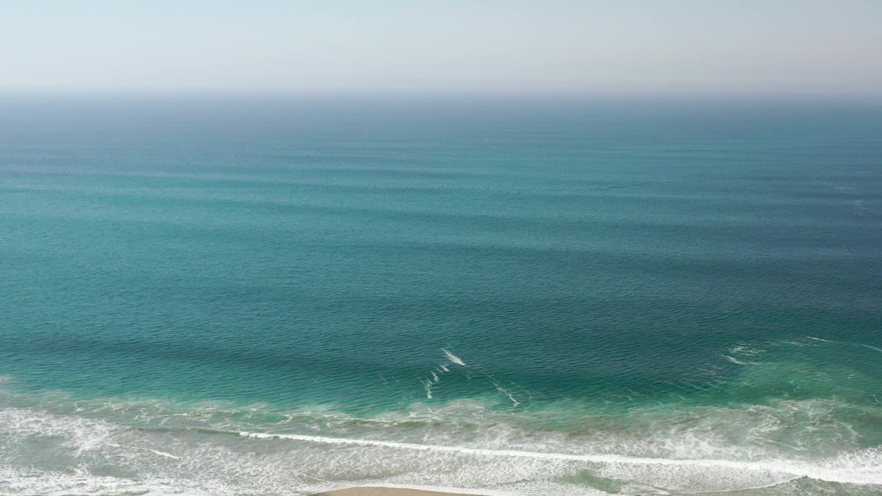 Hazy green-blue ocean with white surf along a sandy shore.