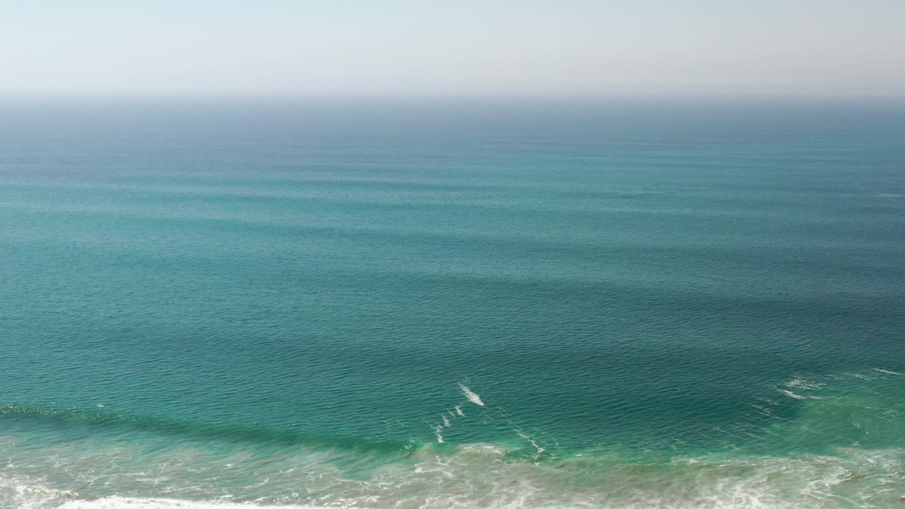Wide Los Cabos ocean view with turquoise water bands, faint white surf near the bottom edge, a pale horizon, and no visible people.