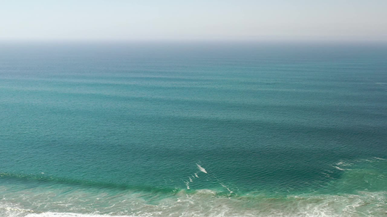 Los Cabos sea surface fills the frame with soft blue-green bands, low white breaking waves along the lower edge, and no visible people.