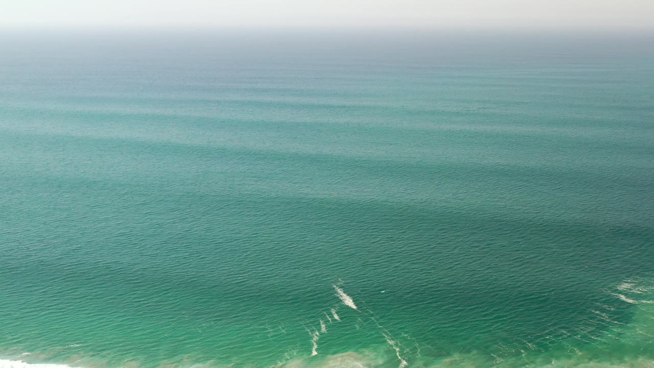 Aerial view of turquoise-green water in Los Cabos, Mexico, with faint white wave breaks lower in frame and no visible people.