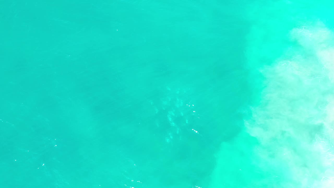 Overhead view of turquoise-green seawater in Los Cabos, Mexico, with a pale foamy wave band on the right and no visible people.
