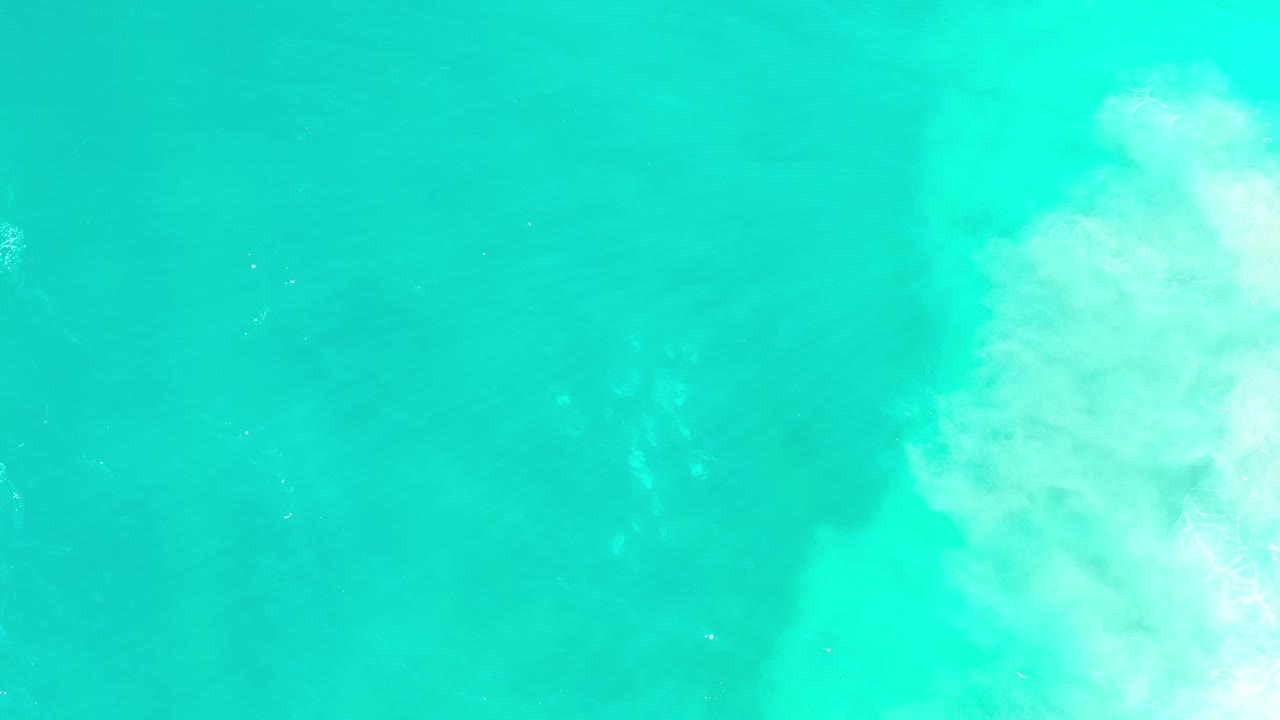 Later overhead frame from Los Cabos, Mexico, with turquoise water filling most of the view, denser white wave wash at the right edge, and no visible people.