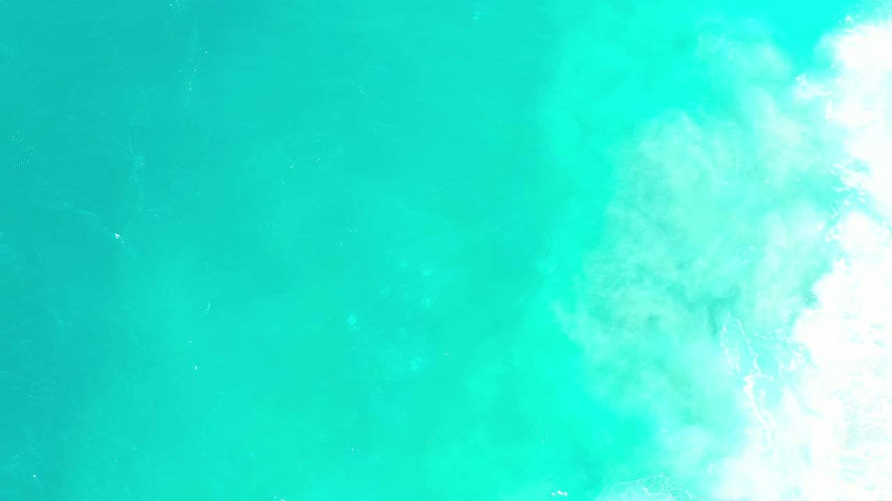 A straight-down ocean view in Los Cabos shows blue-green water with pale foam concentrated near the upper right and no visible people.