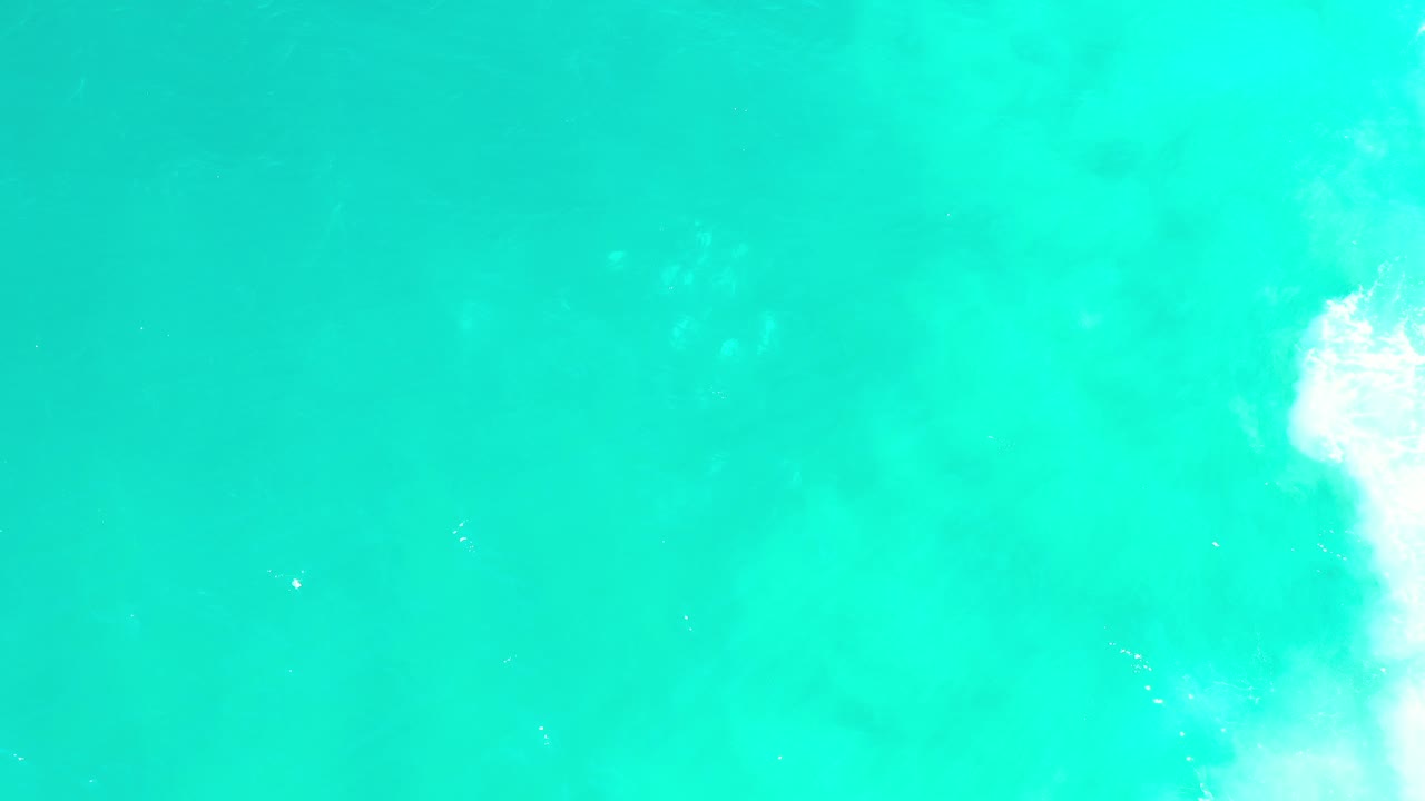 Top-down view of turquoise ocean water in Los Cabos, Mexico, with pale underwater patches and a strip of white surf along the right edge, no visible people.