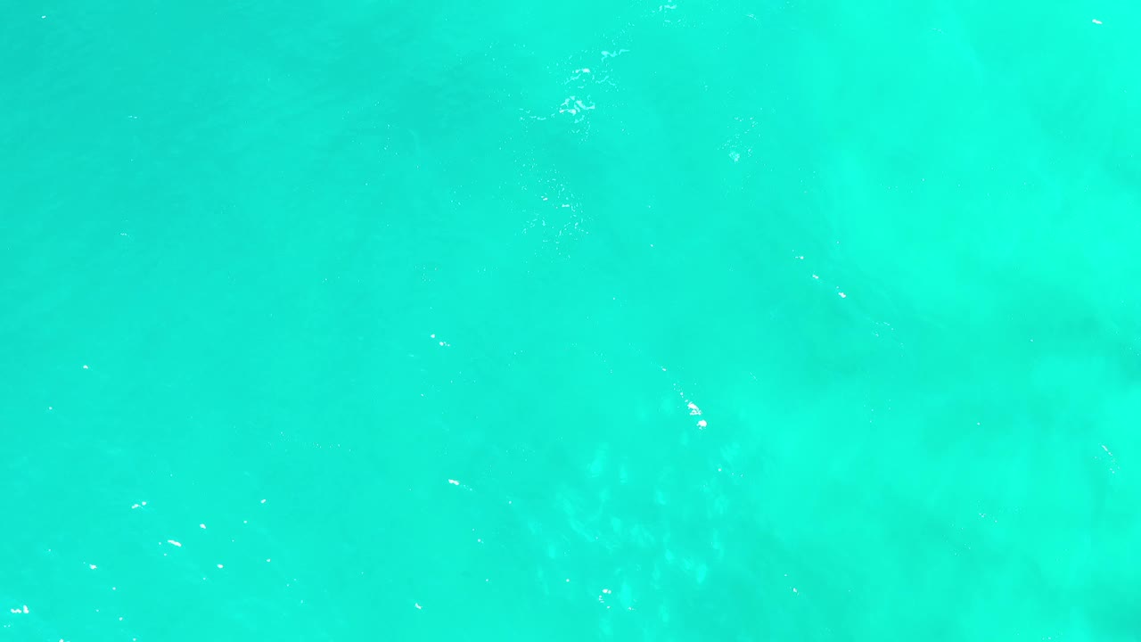 Green-blue water fills the frame from above in Los Cabos, Mexico, with small bright flecks on the surface and no visible people.
