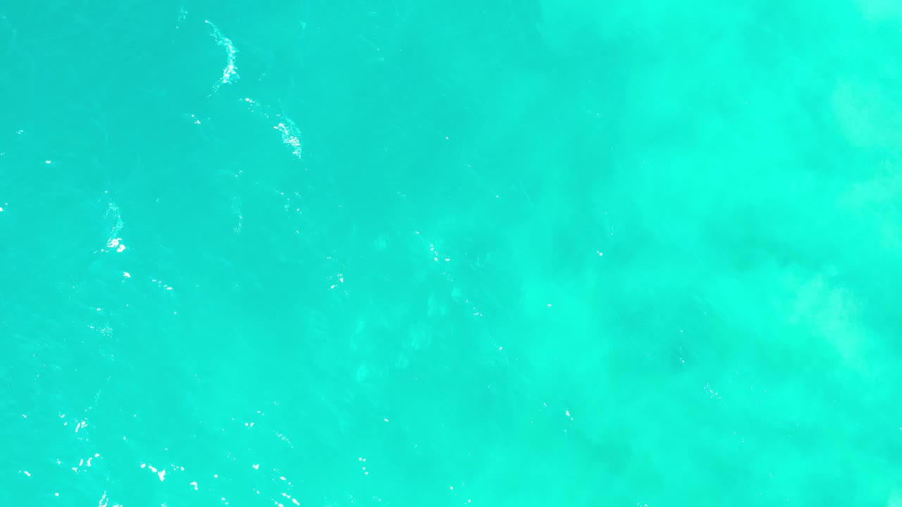 Top-down Los Cabos water view with turquoise color, cloudy darker shapes below the surface, scattered sparkle highlights, and no visible people.