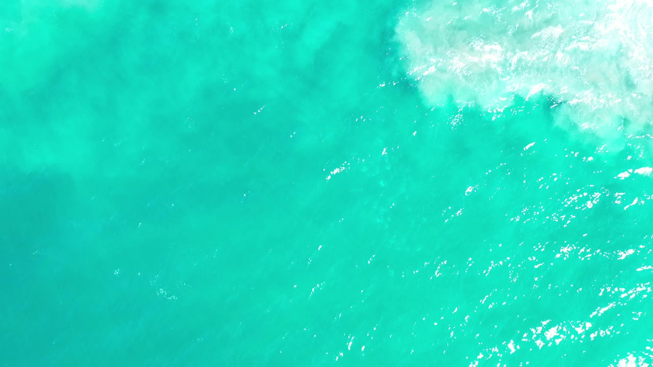 Top-down view of turquoise green water in Los Cabos, Mexico, with a patch of white surf entering at the upper right and no visible people.