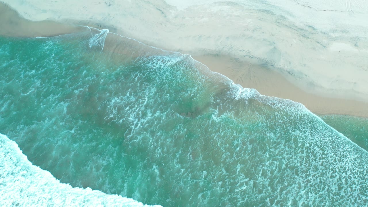 Overhead view of turquoise surf washing onto pale sand in Los Cabos, Mexico, with no visible people.