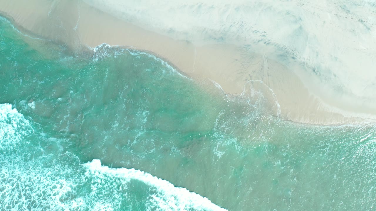 Overhead view in Los Cabos, Mexico, with turquoise water and white foam pushing toward pale sand, no visible people.