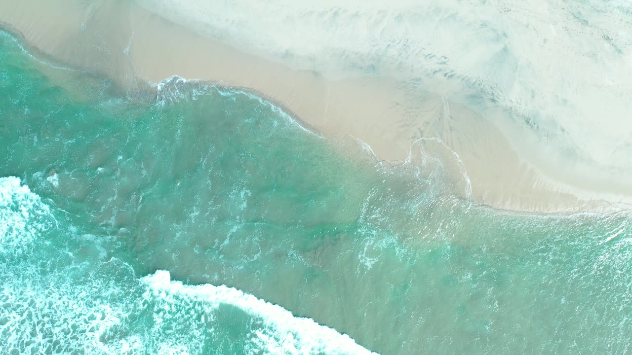 Aerial Los Cabos shoreline frame showing clear green-blue water, a curling foam band, and smooth wet sand, no visible people.