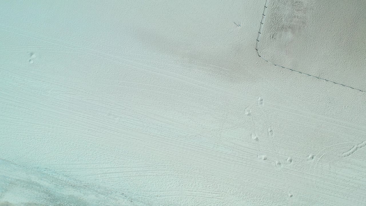 Top-down view of pale beach sand with faint footprints and fencing.