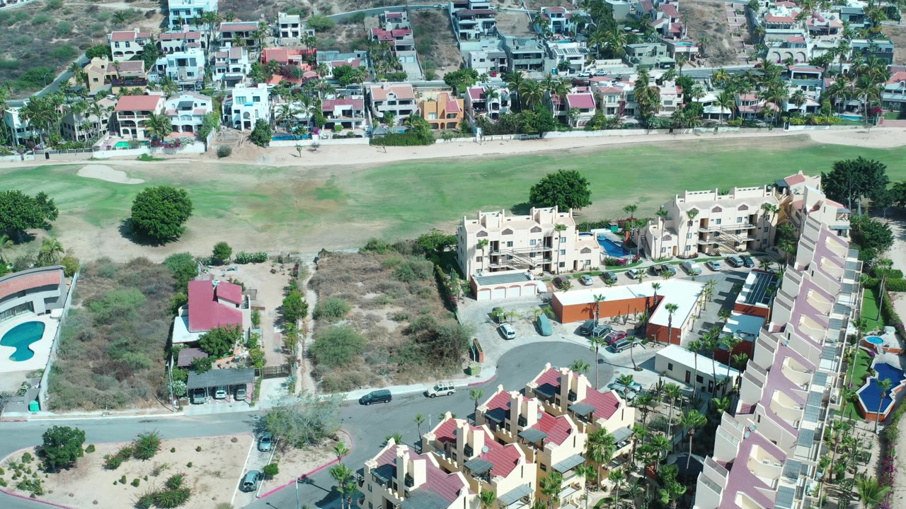 High aerial frame over Los Cabos, Mexico showing cream apartment buildings, red roofs, a road with cars, dry lots, and a green field in the background, with no visible people.
