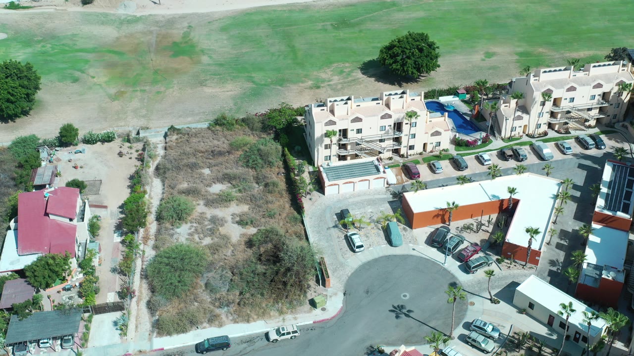 Overhead view of pale buildings, parked cars, dry lots, and open green ground.