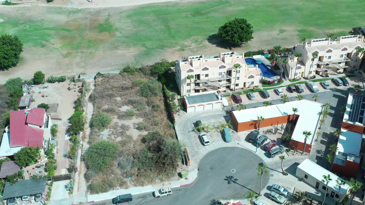 Aerial frame in Los Cabos with a cream building complex, parked cars, dry lot, curved road, and no visible people.