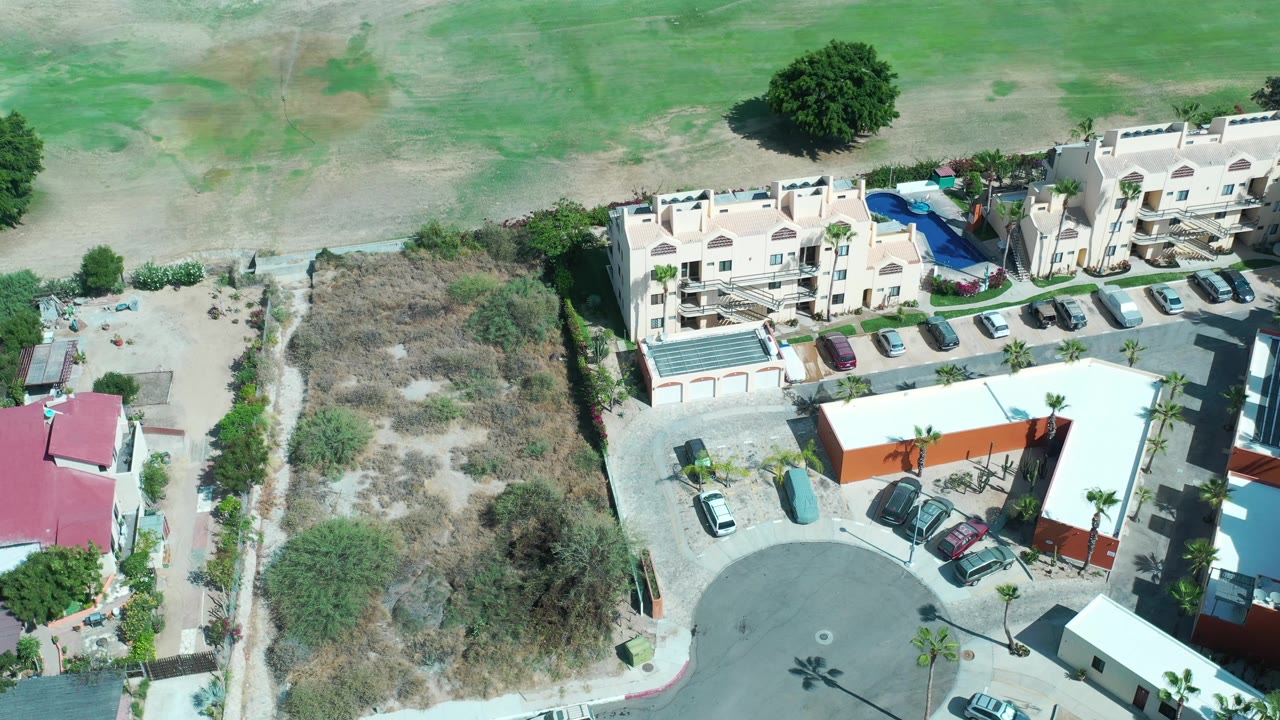 Drone view of cream buildings, a blue pool, parked cars, and arid ground.
