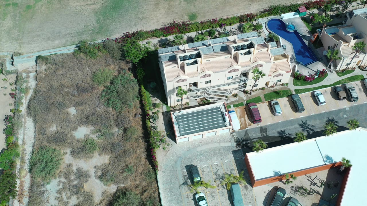 Top-down aerial frame in Los Cabos, Mexico showing a cream hotel complex with parked cars, a blue pool area, palm landscaping, and dry scrub land, with no visible people.