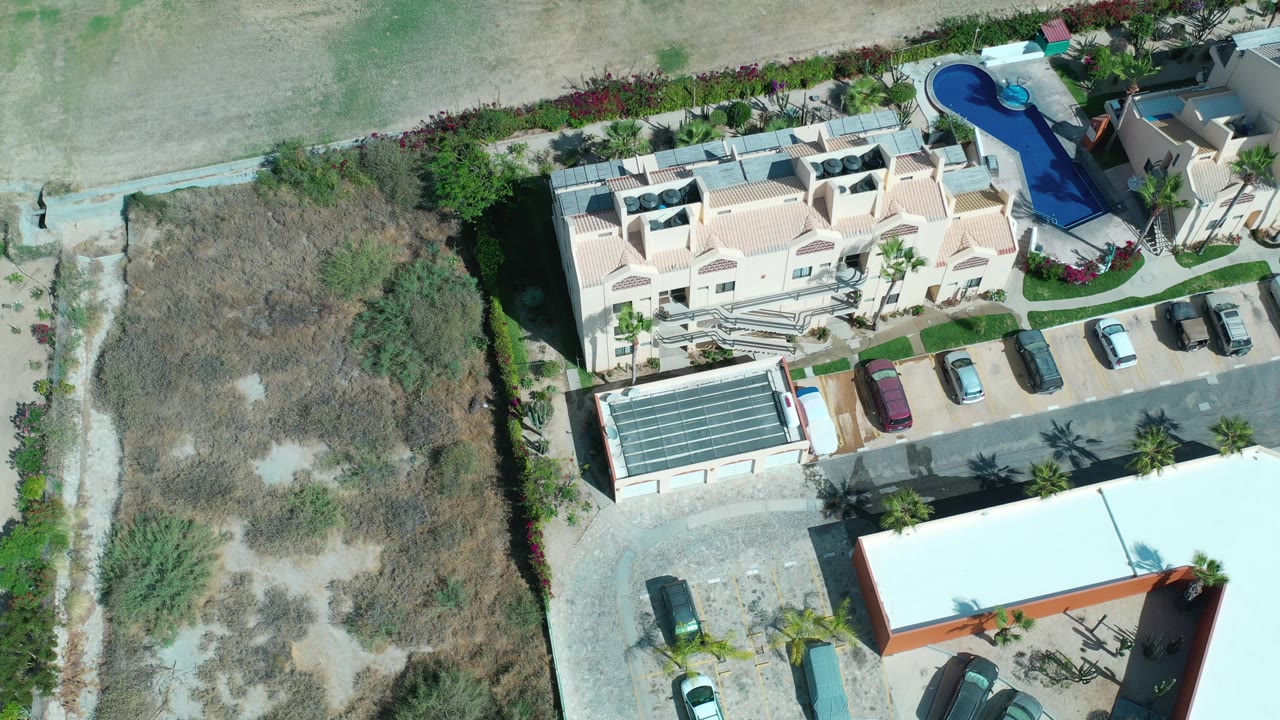 Overhead Los Cabos frame with the hotel roof centered, several parked cars along the driveway, a curved blue pool at the upper right, and no visible people.
