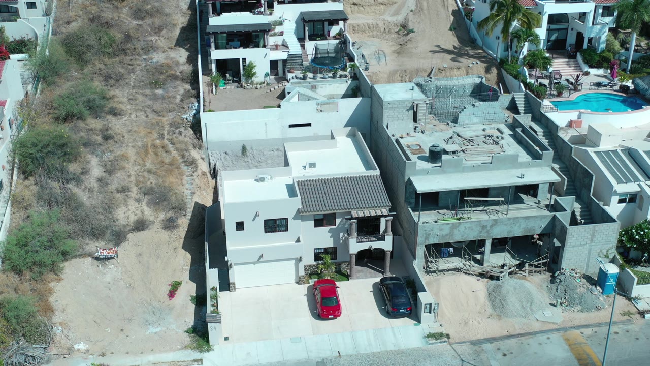 Aerial frame from Los Cabos, Mexico showing white modern homes beside a dry hillside, a red car and dark car in the driveway, construction debris, and no visible people.