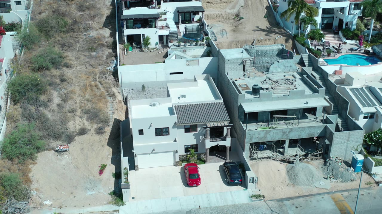 Aerial frame from Los Cabos, Mexico showing the same hillside homes with the unfinished gray structure centered, parked cars below, a blue portable toilet at right, and no visible people.