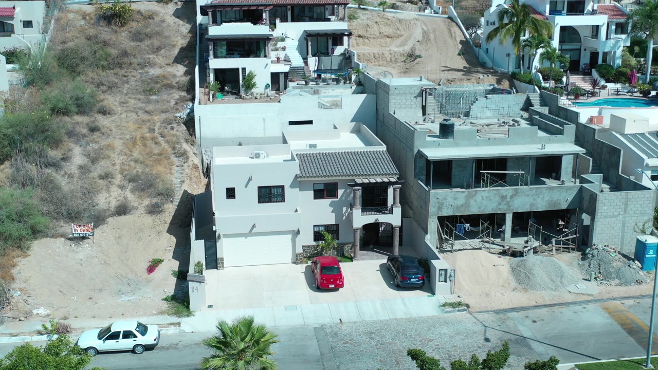 Drone view of hillside homes and a partially built concrete house beside a street.