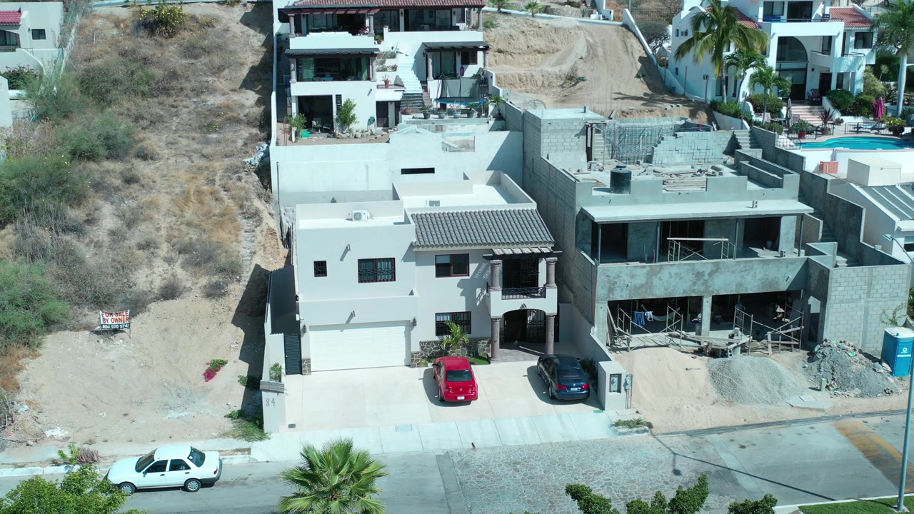 A slightly shifted aerial frame in Los Cabos, Mexico, keeps the red and dark parked cars centered below modern homes, with the dry slope and neighboring houses behind and no visible people.