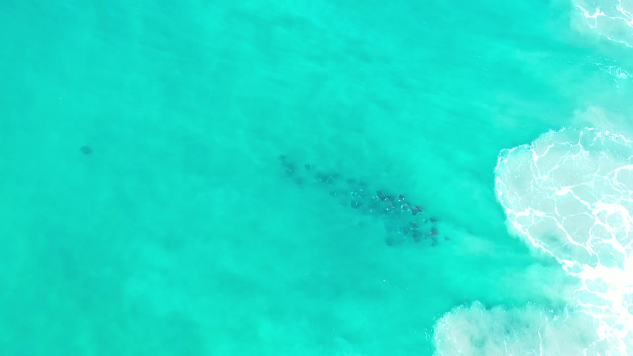 Overhead view of turquoise-green ocean near Los Cabos, Mexico, with a dark submerged patch left of center, white surf on the right edge, and no visible people.
