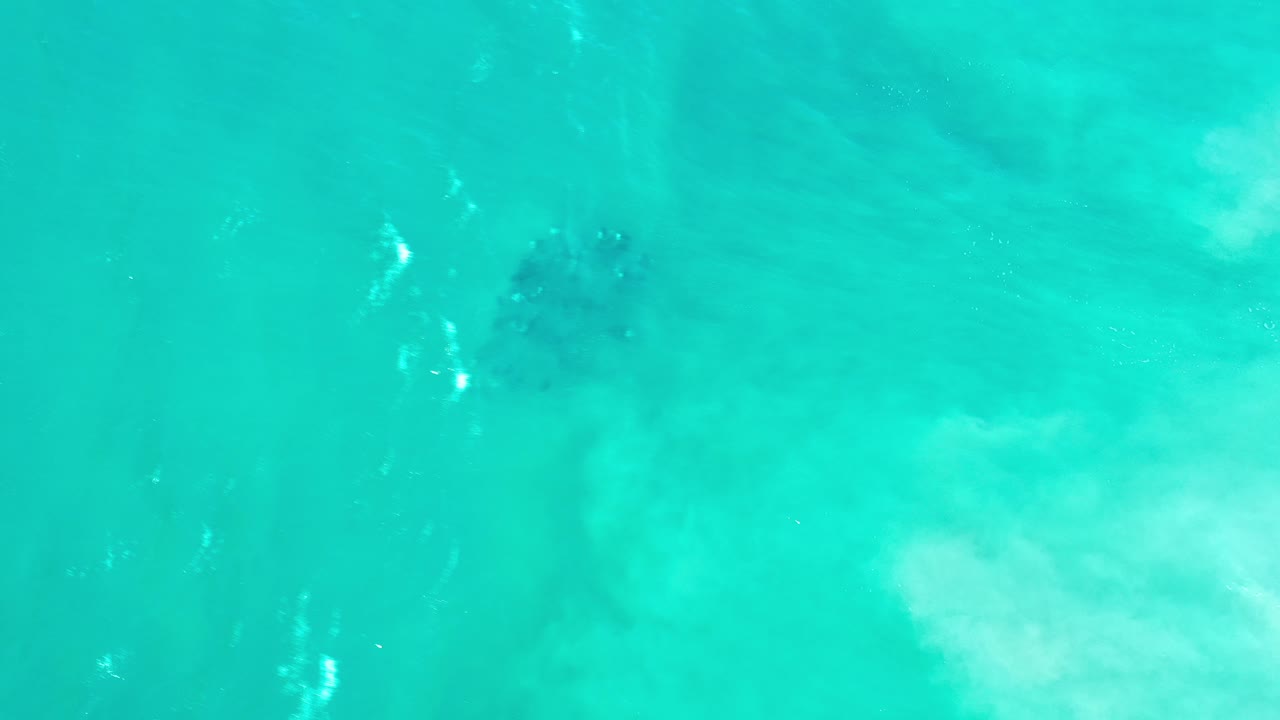 Overhead view of turquoise coastal water in Los Cabos, Mexico, with a dark submerged rock patch near the upper area and no visible people.