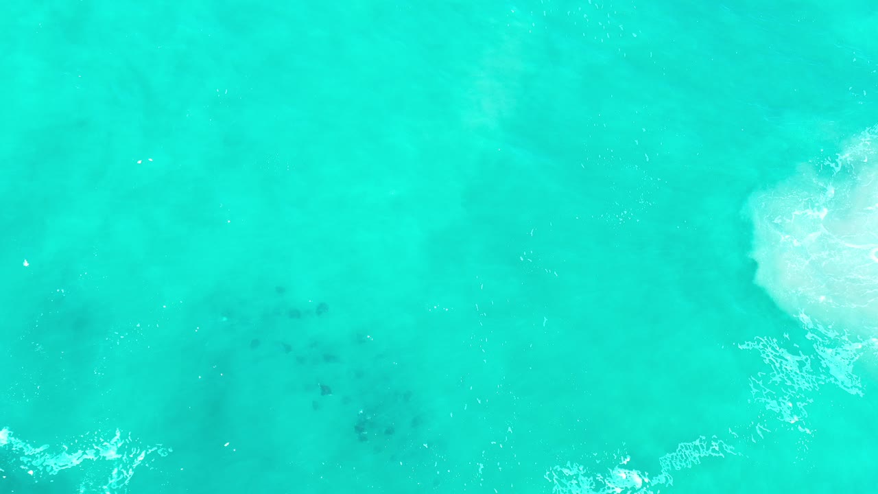 Top-down view of turquoise ocean in Los Cabos, Mexico, with faint darker shapes beneath the water, scattered white foam, and no visible people.